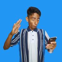 jung asiatisch Mann überrascht suchen beim Clever Telefon im links Hand halten isoliert Blau Hintergrund foto