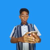 jung asiatisch Mann überrascht suchen beim Clever Telefon, tragen bunt T-Shirt, isoliert Blau Hintergrund. foto