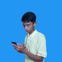 jung asiatisch Mann überrascht suchen beim Clever Telefon, tragen Gelb T-Shirt, isoliert Blau Hintergrund. foto