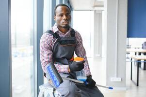 afrikanisch männlich Fachmann Reinigung Bedienung Arbeiter im Overall reinigt das Fenster und Geschäft Fenster von ein Geschäft mit Besondere Ausrüstung foto