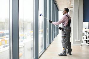 Arbeiter von Reinigung Organisation vorsichtig und vorsichtig reibt groß Fenster von das Büro Raum. ein ernst Afroamerikaner im Blau Overall Tücher das doppelt verglast Fenster im das Büro foto