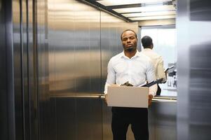 ein jung afrikanisch Mann gefeuert von Arbeit ist Stehen im das Aufzug mit ein Box von Dinge foto