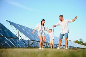 ein breit Schuss von ein glücklich Familie Stehen zusammen und lächelnd beim Kamera mit ein groß Solar- Panel im Hintergrund foto