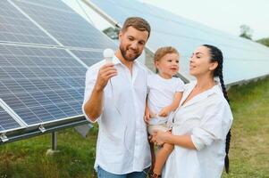 Solar- Energie Konzept. ein jung, glücklich Familie ist Stehen in der Nähe von Solar- Paneele und halten ein elektrisch Licht Birne. foto