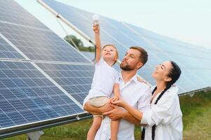 Solar- Energie Konzept. ein jung, glücklich Familie ist Stehen in der Nähe von Solar- Paneele und halten ein elektrisch Licht Birne. foto