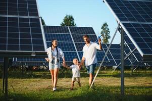 ein breit Schuss von ein glücklich Familie Stehen zusammen und lächelnd beim Kamera mit ein groß Solar- Panel im Hintergrund foto