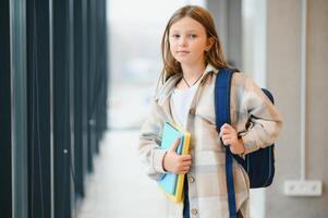 wenig schön Schule Mädchen Stehen unter Gang beim Schule, halten Anmerkungen beim Hände. komisch und glücklich Mädchen lächelnd beim Kamera, ruhen nach Lektionen auf primär Schule foto