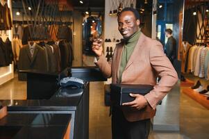 glücklich afrikanisch Käufer halten Kisten von Neu Kleider oder Schuhe und bezahlen mit Anerkennung Karte foto