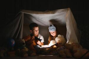 wenig Kinder miteinbeziehen im lesen tolle Buch. Sie Lügen im nett Spielzeug Zelt im Spielzimmer. Junge halten Taschenlampe im Hand. foto