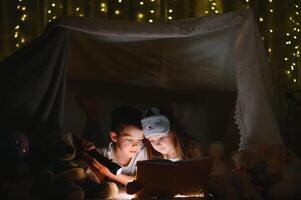 glücklich Familie Kinder lesen ein Buch mit ein Taschenlampe im ein Zelt beim heim. foto