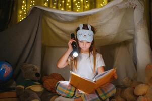 glücklich Kind Mädchen Lachen und lesen Buch im dunkel im ein Zelt beim heim. foto