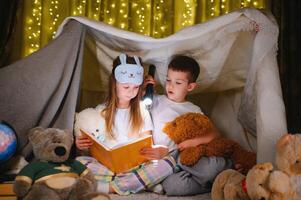 zwei Kinder mit Taschenlampe lesen ein Buch unter ein Decke wie ein Zelt. foto