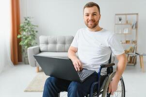 Porträt von lächelnd deaktiviert männlich Sitzung im Rollstuhl und Arbeiten auf Laptop von heim. jung Arbeiter mit Besondere braucht. Freiberufler und Menschen mit Behinderungen Konzept. foto
