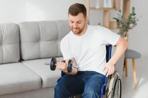 Sportler Sitzung im Rollstuhl und Ausstrecken Waffen mit Hanteln während Rehabilitation Übung im modern medizinisch Center. Mann Sitzung auf Rollstuhl beim heim. foto