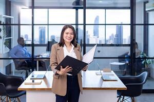 Porträt von asiatisch Geschäft Vorsitzender Frau ist Stehen im das Büro beim das Tabelle mit dokumentieren Datei und zeigen Statistik Diagramm zeigen jährlich Bericht und Wolkenkratzer Hintergrund foto