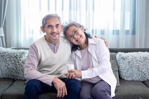 Paar von gesund Ehe asiatisch Senior Vater und Mutter Sitzung zusammen mit Glas von Milch und glücklich Lächeln auf gemütlich Couch im Pensionierung Zuhause während Winter zum ältere Pflege zu Ausgaben wertvoll Zeit foto