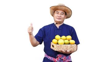gut aussehend asiatisch Mann Farmer trägt Hut, Blau Shirt, hält Korb von organisch Orange Früchte, Daumen hoch, isoliert auf Weiß Hintergrund. Konzept, Landwirtschaft Beruf, produzieren Pflanzen zu Markt. foto