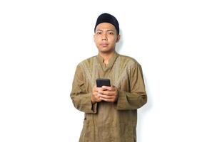 verwirrt asiatisch Muslim Mann tragen islamisch Kleider mit Smartphone, suchen beim Kamera, isoliert auf Weiß Hintergrund foto