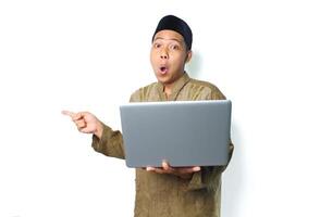 überrascht asiatisch Muslim Mann zeigen zu leeren Raum mit halten Laptop, suchen beim Kamera, isoliert auf Weiß Hintergrund foto