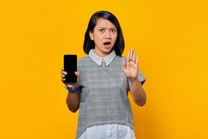 Porträt einer überraschten jungen asiatischen Frau mit offenem Mund, die ein Smartphone mit leerem Bildschirm zeigt und eine Stoppgeste mit Handflächen macht foto