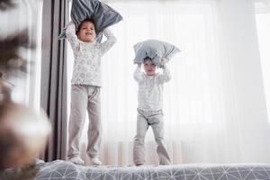 Kinder, die im Bett der Eltern spielen. Kinder wachen im sonnigen weißen Schlafzimmer auf. Junge und Mädchen spielen in passenden Pyjamas. Familienmorgen foto