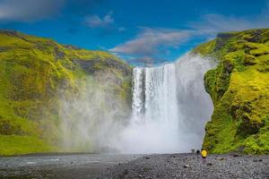 mächtiger und berühmter skogafoss-Wasserfall mit einer einsamen stehenden Person in orangefarbener Jacke, beim Wandern in Island, im Sommer, malerischer dramatischer Aussicht bei sonnigem Tag und blauem Himmel. foto