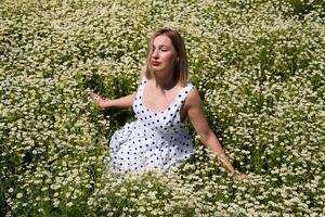 ein Frau im ein Weiß Kleid mit Polka Punkte ist auf ein Lichtung mit Gänseblümchen. Blühen Gänseblümchen foto