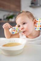 süß Baby Mädchen Kleinkind Sitzung im das hoch Stuhl und Essen ihr Mittagessen Suppe beim Zuhause Küche. foto