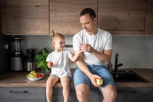 süß wenig Mädchen und ihr gut aussehend Papa sind Essen Obst im modern Küche. gesund Essen. foto