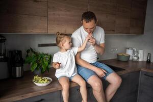 süß wenig Mädchen und ihr gut aussehend Papa sind Essen Obst im modern Küche. gesund Essen. foto