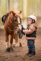 Porträt von wenig Mädchen im schützend Jacke und Helm mit ihr braun Pony Vor Reiten Lektion foto