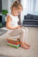 wenig Mädchen sitzt auf ein Stapel von Kinder- Märchen Bücher und Theaterstücke das App auf ihr Smartphone foto