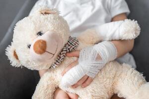 wenig Mädchen mit gebrochen Finger hält Teddy Bär mit ein verbunden Pfote beim das Arzt geplanter Termin im das Krankenhaus foto