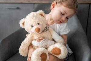 wenig Mädchen mit gebrochen Finger hält Teddy Bär mit ein verbunden Pfote beim das Arzt geplanter Termin im das Krankenhaus foto