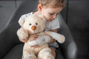 wenig Mädchen mit gebrochen Finger hält Teddy Bär mit ein verbunden Pfote beim das Arzt geplanter Termin im das Krankenhaus foto