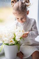 wenig Mädchen Sitzung durch Fenster mit Tulpe Blumen Strauß. glücklich Kind, drinnen. Mutter Tag, Valentinstag Tag oder Geburtstag Konzept. foto