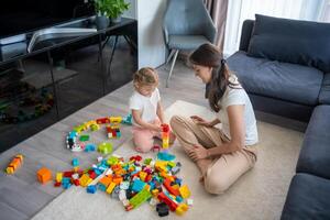 wenig Mädchen abspielen mit Konstrukteur Spielzeug auf Fußboden im Zuhause mit Mama oder Frau Babysitter, lehrreich Spiel, Familie beim Zuhause verbringen Freizeit Aktivitäten Zeit zusammen Konzept foto