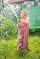 glücklich Lachen Kind Mädchen 2-3 Jahr alt tragen wasserdicht Kleider haben ein Spaß mit Wasser Gewehr auf Zuhause Hinterhof foto