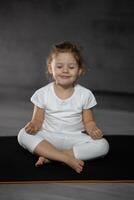 drei Jahre alt wenig Mädchen meditieren im ein Lotus Pose auf ein grau Hintergrund im dunkel Zimmer. hoch Qualität Foto