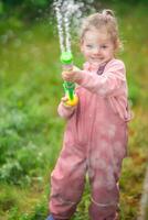 glücklich Lachen Kind Mädchen 2-3 Jahr alt tragen wasserdicht Kleider haben ein Spaß mit Wasser Gewehr auf Zuhause Hinterhof foto