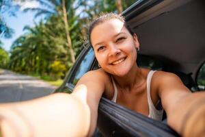 jung Frau mit dunkel Haar stecken ihr Hände aus von das Taxi von ein Auto und machen Selfie auf das Straße, Konzept von Auto Reise. hoch Qualität Foto