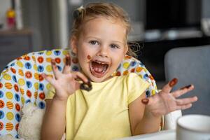 wenig Mädchen mit blond Haar Essen hausgemacht Schokolade und zeigen Mund und schmutzig Hände mit Flecken von Schokolade im Zuhause Küche foto