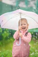 glücklich Lachen Kind Mädchen 2-3 Jahr alt tragen wasserdicht Kleider und halten Rosa Regenschirm haben ein Spaß auf Zuhause Hinterhof im regnerisch Tag foto