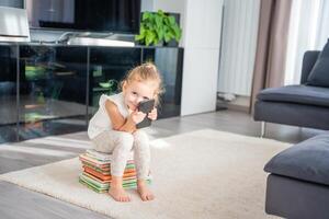 lächelnd wenig Mädchen sitzt auf ein Stapel von Kinder- Bücher und Verwendet ihr Smartphone foto