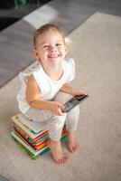 lächelnd wenig Mädchen sitzt auf Stapel von Kinder- Märchen Bücher mit Smartphone im ihr Hände foto