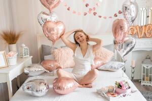 schön jung Mädchen beim Zuhause auf das Bett im das Morgen genießen Valentinsgrüße Tag Feier mit Herz geformt Luftballons. hoch Qualität Foto