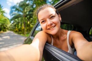 glücklich jung Frau mit dunkel Haar stecken ihr Hände aus von das Taxi von ein Auto und machen Selfie auf das Straße, Konzept von Auto Reise. hoch Qualität Foto