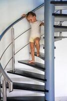 Porträt von wenig Mädchen gehen Nieder das Treppe beim modern heim, Kind Klettern Spiral- Treppe. gefährlich Situation beim heim. Kind Sicherheit Konzept. foto