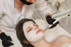 weiblich Fachmann Kosmetikerin Hände bewirbt sich Maske zu Patienten Gesicht zum Gesichts- Haut Pflege. weiblich Klient entspannend mit geschlossen Augen und genießen Spa foto