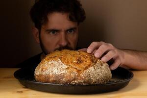 attraktiv jung kaukasisch Koch posieren mit Weiß Sauerteig Brot. das Sauerteig Brot ist das zentral Protagonist von das Szene, Stehen aus mit schön golden Töne gegen das dunkel Hintergrund. foto